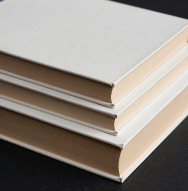 Close-up of a stack of hardcover books with clean, minimalist covers in soft off-white and muted golden tan tones, arranged on a deep charcoal black surface. High-fashion editorial photography style in a South American / Brazilian setting.