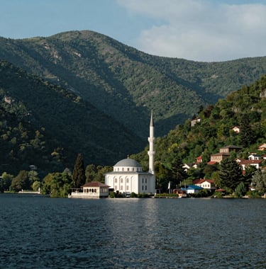 A scenic view of Uzungol lake in Trabzon with the iconic white mosque at its edge. Lush green mountains rise sharply around the calm water under a soft sky blue sky. Eastern European / Turkish architecture and nature photography.