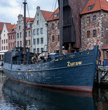 A detailed photograph of the historic wooden Port Crane 'Zuraw' in Gdansk. The weathered timber is a deep Muted Slate Blue under soft daylight. The surrounding stone architecture is Soft Steel Grey, and the scene is reflected in the calm, Dark Navy waters of the Motlawa River.