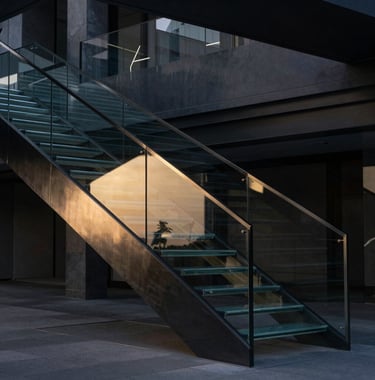 A cinematic, architectural shot of a modern glass staircase in a darkened building, reflecting a subtle gold light. International business aesthetic, sharp and precise.