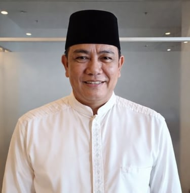 Smiling man wearing a traditional black songkok hat and white baju melayu shirt in an office setting.
