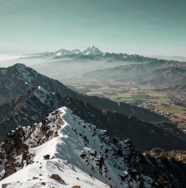 A majestic panorama shot from the top of Phulchoki Peak showing the snowy Himalayan mountains on one side and the vast Terai plains on the other. Colors include dark slate grey peaks and pale mist green skies.