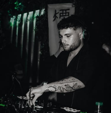 Professional DJ with arm tattoos performing a live set at a dark nightclub with green lighting.