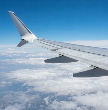 A close-up photograph of a luxury airplane wing flying above a sea of white clouds against a vibrant, clear blue sky. The image represents the excitement and freedom of international travel.