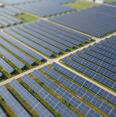 An aerial view of a solar farm integrated into a green landscape. The shot is bright, professional, and clean, showing the intersection of technology and nature. Strong presence of #7BA6A8 and #E8F2F3 tones.