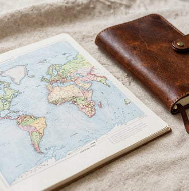 An intimate, editorial-style photo of a hand-sketched travel map and a leather-bound journal on a sand-colored linen cloth. Soft afternoon light. Global / Discerning English-speaking aesthetic.