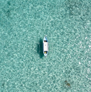 An aerial photography shot of a single small boat floating on crystal clear, light cyan water in the South American Caribbean. The composition is clean and minimalist, showcasing the depth and clarity of the ocean.