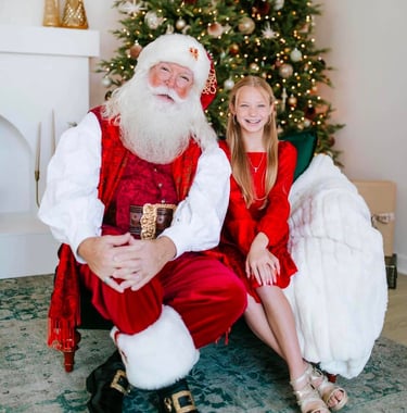 Smiling girl sitting with Santa Claus in front of a decorated Christmas tree with lights.