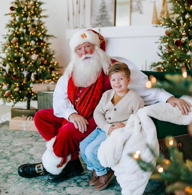 A smiling young boy sits with Santa Claus in a festive room with decorated Christmas trees.