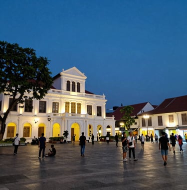 An evocative evening photo of the Batavia Old Town square in Jakarta, showing the colonial architecture lit up, with people enjoying the vibrant atmosphere in a Southeast Asian / Indonesian urban setting.