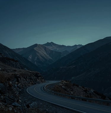 A cinematic, wide-angle landscape shot of a quiet mountain road winding through peaks. The lighting is dusk, casting professional deep blue (#1E2B38) and muted teal (#4A7B8E) shadows over the vista. Minimalist and grand.