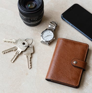 A flat lay of professional real estate tools including a set of luxury home keys, a high-end watch, a leather folio, and a smartphone on a polished beige marble surface. Middle Eastern / Gulf professional style.