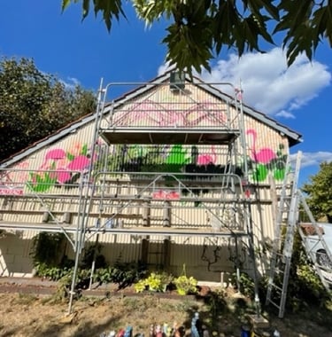 une grange en taule avec un échafaudage et une fresque avec des flamands roses et des plantes