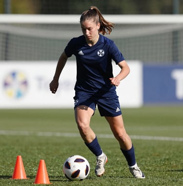 A focused European teenage girl practicing dribbling around cones, displaying high intensity, dressed in professional training gear with Total Football Academy logo details, bright daylight.