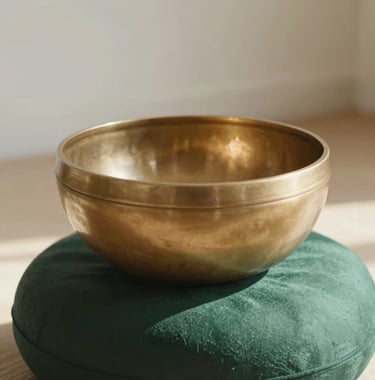 A close-up photograph of a brass singing bowl on a dark forest green cushion. Sunlight catches the metal, and the background is a soft off-white, suggesting a meditative Western European / French holistic space.