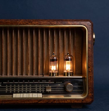 Close-up photograph of a vintage 1930s wooden tabletop radio receiver, with warm glowing vacuum tubes visible through the rear grill. Set against a dark slate navy blue background, styled for a scholarly Eastern European digital archive.