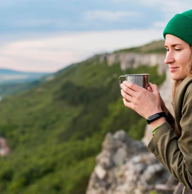 Uma mulher jovem segurando uma xícara admirando o horizonte no alto de uma montanha
