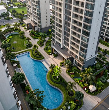A high-angle architectural photograph of a premium residential complex featuring beautifully landscaped green gardens, swimming pools, and modern walking paths in a South Asian urban setting.