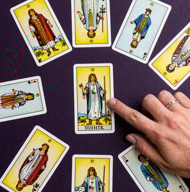 A top-down view of a spread of Tarot cards on a dark purple surface, with a delicate hand wearing a silver ring pointing to a card, South American style.