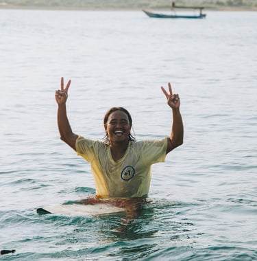 Smiling local surfer in tropical water making peace signs while sitting on a surfboard.