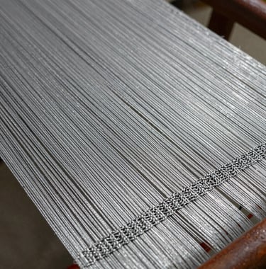 Close-up macro photography of silver metallic silk threads being woven into a complex pattern, showcasing the intricate detail and luxury quality of Surat textile engineering.