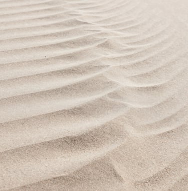A close-up shot of wind-blown sand forming patterns that mimic ocean waves. The colors range from deep #DDCAB8 to light #FAF8F5, emphasizing texture and natural design.