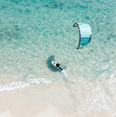 An artistic, top-down view of a kitesurfing board partially submerged in crystal clear turquoise water, casting a soft shadow on the white sand #FAF8F5 below. The composition is minimal and highlights the movement and energy of the ocean.