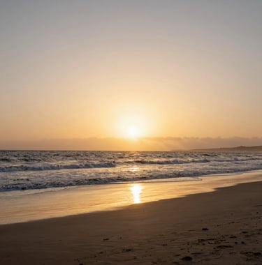 A landscape shot of the beach in Agadir at sunset. The sky is a mix of golden sand tan and soft cream. The Atlantic waves are gentle, and the sand reflects the warm evening light.