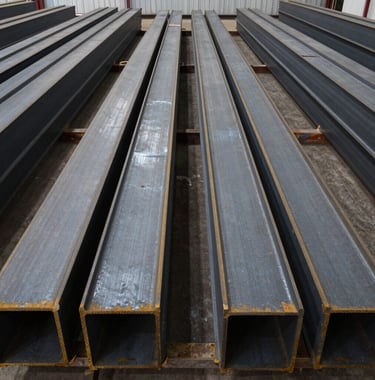 Wide shot of rows of structural steel beams ready for shipment in a large North American / US warehouse. The symmetry and scale of the dark slate steel beams convey leadership and massive production capacity.