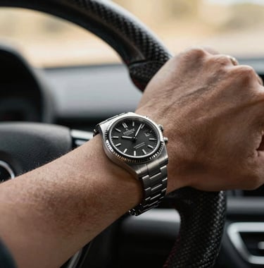 A close-up shot of a luxury watch on a driver's wrist holding a carbon fiber steering wheel, with soft champagne gold bokeh in the background.
