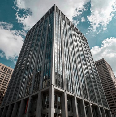Wide architectural shot of a sleek, modern commercial building in a major North American financial district. The facade is glass and steel, reflecting the crisp white clouds and vibrant teal sky. Premium, authoritative, and stable atmosphere.