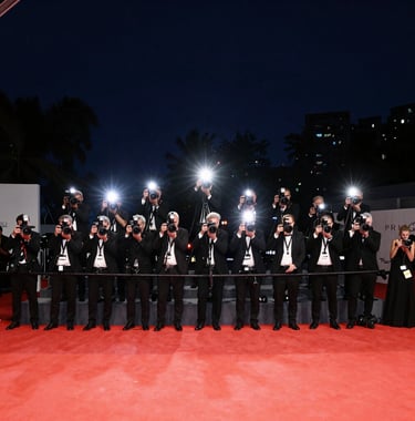 A wide-angle shot of a premiere red carpet event at night. The flashes of cameras create a sparkling light effect against the dark navy night sky. A sense of prestige, stardom, and modern cinematic luxury.