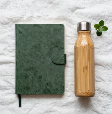 A flat lay of travel essentials: a dusty leaf green notebook, a bamboo water flask, and a small plant on soft misty white linen. Clean and professional composition.