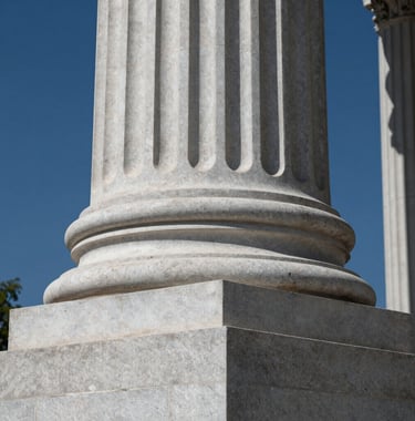 A low-angle shot of a sturdy stone pillar or pedestal, representing a firm foundation for institutional work. The lighting is strong and clear, highlighting the solid texture of the stone. Colors integrated: #94A7B7 and #2B4E6B.