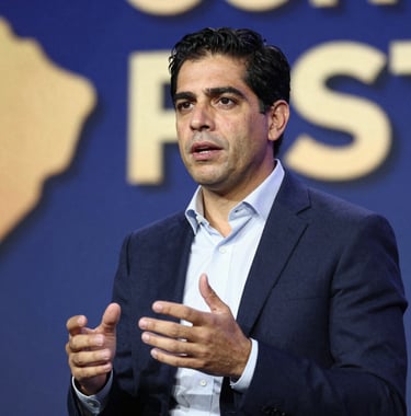 A candid shot of Adrián Andrade Castro speaking at a business conference in South America, looking authoritative yet approachable. The background shows blurred blue and gold stage lighting.