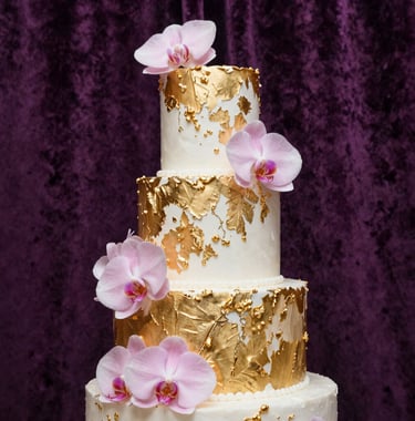 A close-up of a three-tier wedding cake decorated with real gold leaf and soft pink orchids, set against a dark purple velvet background with soft backlighting.