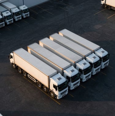A luxury aerial view of a massive logistics hub with several white transport trucks perfectly aligned against a dark black asphalt ground, with long evening shadows and subtle gold highlights.