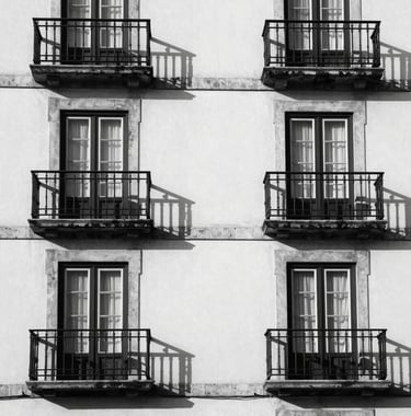 A minimalist architectural detail of a classic Portuguese building facade with clean white stone and dark metal balconies. High contrast between shadows and light, capturing a sense of heritage and quality. Black and white photography.