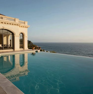 A wide shot of a luxury villa infinity pool overlooking the ocean in a Western / International setting. The water in the pool is deep charcoal teal, perfectly reflecting the elegant beige architecture of the terrace.