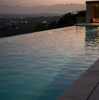 A close-up of a crystal-clear infinity pool at dusk. The water reflects the dark charcoal tones of the evening sky and the warm, champagne taupe glow of outdoor lighting. The composition is sleek and tranquil, emphasizing luxury outdoor living.