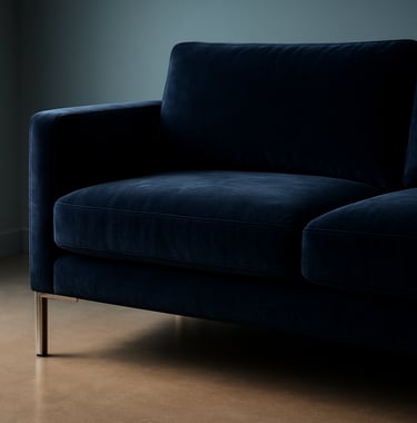 A close-up photograph of a luxury dark navy velvet sofa with polished steel legs, placed against a pale blue wall in a modern Indian home. Soft evening lighting highlights the premium fabric texture and sophisticated design.