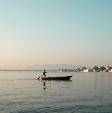 A wide landscape of a serene lake in Udaipur at dawn, a lonely South Asian / Indian boatman paddling across the water, soft pastel colors of sand and light aqua in the sky.