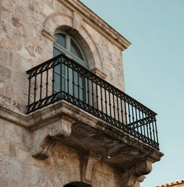 Architectural detail of a classic wrought-iron balcony and stone arch in Pézenas, set against a soft blue-green sky, capturing the authentic Southern European charm.