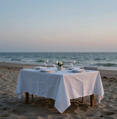 A minimalist, beautifully set dinner table on a private beach under a twilight sky, with soft blue and white linens.