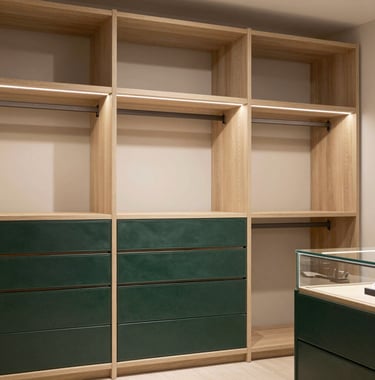 A luxury walk-in wardrobe with soft off-white ambient lighting. The shelving is finished in a warm sandy beige wood, with deep forest green leather accents on the drawers and glass display cases.