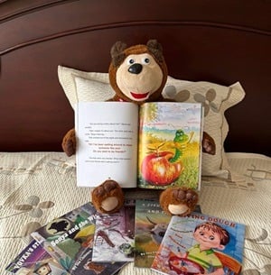 A smiling teddy bear holding an open picture book with four other books on the bedspread.