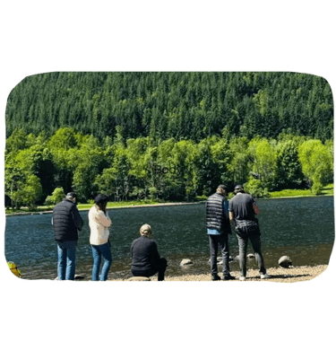 a group of people standing beside a loch with trees across the water