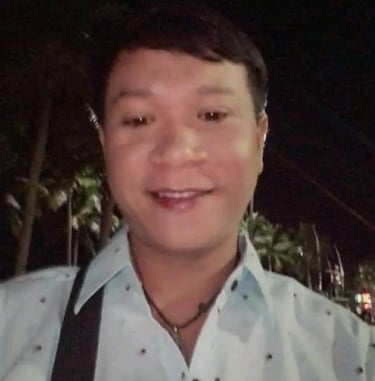 Man smiling outdoors at night in Pattaya, Thailand, wearing a light shirt and shoulder bag.