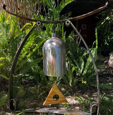 A metal outdoor garden bell hanging from a rustic iron stand in a lush backyard landscape with green ferns.
