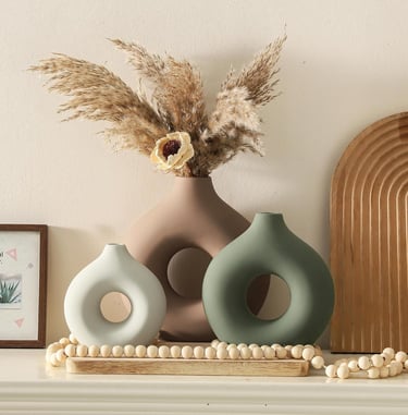 Three modern ceramic donut vases in neutral tones with dried pampas grass on a mantel.